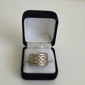May include: A silver-toned metal ring featuring a continuous woven knot pattern around the band. The ring is displayed inside a small, rectangular black velvet jewelry box with a white interior lining and a hinged lid that is open.