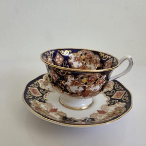 May include: A porcelain teacup and saucer set featuring a deep navy blue and white colour palette. The design includes intricate gold filigree, floral patterns in shades of orange and yellow, and diamond-shaped accents with gold trim on the edges.