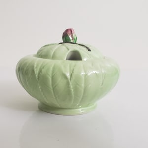 Peut inclure: Un sucrier en céramique vert clair avec un couvercle. Le bol présente un motif de feuilles et un petit bouton de fleur rose et vert sur le dessus. Le bol a une petite base et un trou dans le couvercle.
