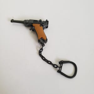 May include: Miniature black and brown pistol keychain with a brown grip. The keychain is attached to a black chain and a black key ring. The pistol is a replica of a Luger.