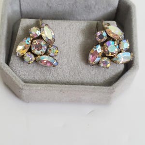 May include: A pair of vintage clip-on earrings with clusters of iridescent, oval, and round rhinestones. The stones display pink, yellow, and blue hues, set in gold-tone metal. The earrings are displayed in a grey velvet jewellery box.