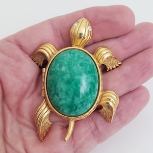 Puede incluir: Broche de tortuga dorada con un gran centro de piedra verde ovalada. La cabeza y las aletas de la tortuga presentan diseños texturizados. Dos pequeños cristales transparentes adornan la cabeza.