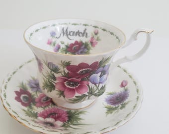May include: A white teacup and saucer with a floral design. The cup has a gold rim and the words "March" printed on the side. The saucer has a matching floral design.