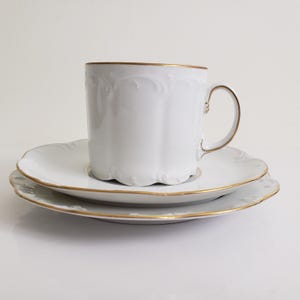 May include: A white porcelain teacup and saucer set with gold edging. The cup has a scalloped rim and a curved handle. The set includes two saucers, also with gold trim. Ideal for serving tea or coffee, the set is elegant.