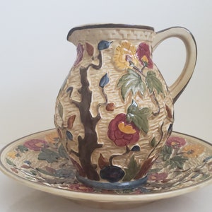 May include: A cream-coloured ceramic jug with a floral design and a matching plate. The jug has a brown tree branch and green leaves with red, yellow, and blue flowers. The plate has a similar floral design.
