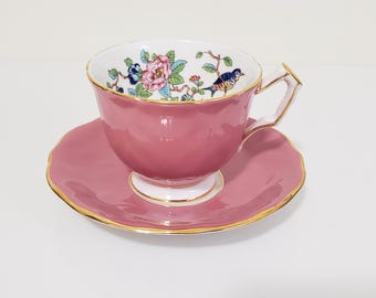 May include: A pink teacup and saucer set with gold trim. The teacup features a floral and bird design inside the cup. The saucer has a scalloped edge. The set is on a white background.