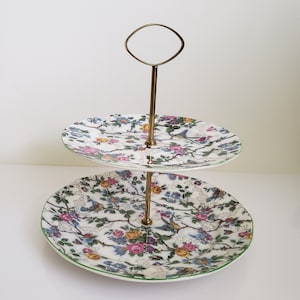 May include: Two-tiered cake stand with a gold metal handle. The stand is decorated with a floral pattern featuring pink, yellow, and blue flowers, green leaves, and blue birds.