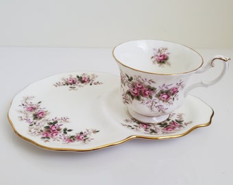 May include: A white teacup and saucer set with a pink floral pattern. The saucer has a scalloped edge and a gold trim. The teacup has a handle and a gold trim around the rim.