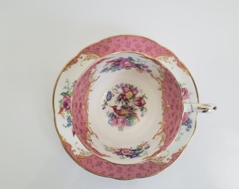 May include: A pink and gold teacup and saucer set with a floral pattern and a bird design. The cup and saucer are both decorated with a delicate floral pattern and a gold trim.