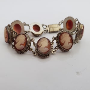 Puede incluir: Un brazalete vintage con camafeos que presenta camafeos ovalados con retratos de perfil en tonos crema y marrón. Los camafeos están engastados en marcos plateados ornamentados y unidos por una delicada cadena. El brazalete se asegura con un cierre.