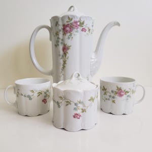 May include: A white porcelain tea set with a coffee pot, sugar bowl, and two mugs. The set is adorned with pink roses and blue floral patterns. The coffee pot has a large handle and a long spout. The set is ideal for serving tea.