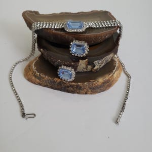 May include: A silver-coloured necklace and matching earrings set. The necklace and earrings feature light blue rectangular centre stones surrounded by clear rhinestones. The jewellery is displayed on a brown and tan stone.