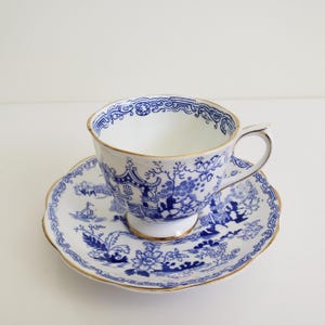 Könnte beinhalten: Weißes Tee-Service mit einer Tasse und Untertasse mit blau-weißem Blumen- und Landschaftsdesign. Die Tasse und die Untertasse haben einen goldenen Rand. Die Tasse hat einen Henkel und steht auf einem kleinen Sockel.