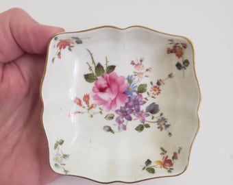 May include: A small, square, white porcelain dish with a scalloped edge and a gold trim. The dish is decorated with a floral pattern of pink, purple, and yellow flowers.