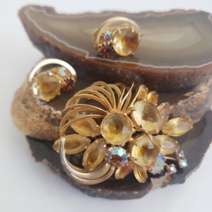 Può includere: Un set di gioielli vintage con una spilla dorata e orecchini abbinati. La spilla ha un design floreale con grandi pietre color ambra e piccoli accenti iridescenti. Anche gli orecchini hanno pietre color ambra. Il set è esposto su una pietra decorativa.