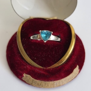 May include: A silver ring featuring a triangular, faceted, bright blue gemstone set in a three-prong mounting. The ring is displayed inside a heart-shaped jewelry box with a deep red velvet interior and a gold-toned metal rim.