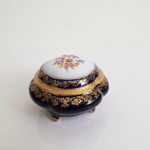 May include: A small, ornate jewellery box with a navy blue base and lid. The lid has a white centre with a floral design in various colours. Gold trim and accents adorn the box, which rests on three small feet.