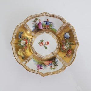 Könnte beinhalten: Eine weiße Porzellansaucerschale mit einem gewellten Rand und goldenen Verzierungen. Die Saucerschale ist mit vier gemalten Szenen von Menschen in einer Landschaft und einem zentralen Blumenmotiv verziert.