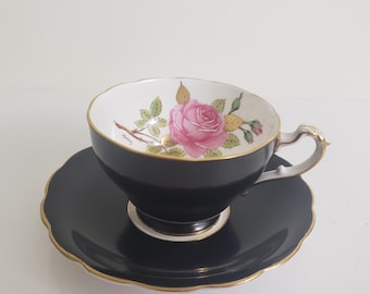 May include: A black teacup and saucer set with gold trim. The teacup features a pink rose design inside. The saucer has a scalloped edge. The handle of the teacup is gold-colored.