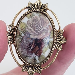 Peut inclure: Une broche ovale en ton or avec un motif floral en relief détaillé. Le centre présente un camée blanc avec une image peinte de deux roses roses.
