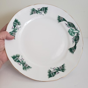 May include: A white ceramic dinner plate with a gold rim and a green and white tartan ribbon design. The ribbon is tied in a bow and has a small sprig of heather flowers.