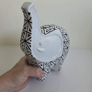 以下が含まれることがあります： 白い陶器製の象の置物で、体全体に黒い幾何学的な三角形の模様が施されています。象は鼻を上げ、耳の部分は滑らかで、光沢のある仕上げになっています。黒い線が繰り返され、三角形を形成するデザインです。