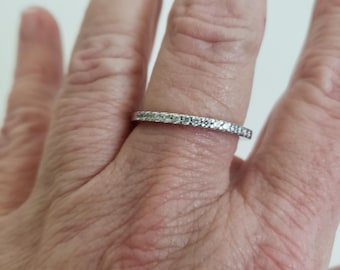 May include: A silver ring with a band of small, clear gemstones. The ring is on a finger, with the hand in the frame. The ring is simple and elegant, with a classic design.