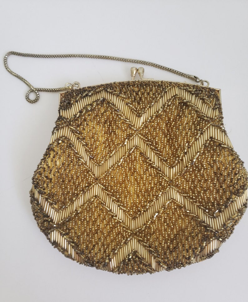 Pu&ograve; includere: Una borsa da sera in perline dorate con un motivo a chevron. La borsa ha una tracolla a catena argentata.