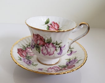 May include: A white teacup and saucer with a floral pattern of pink, purple, and green sweet peas. The cup and saucer have a gold trim.