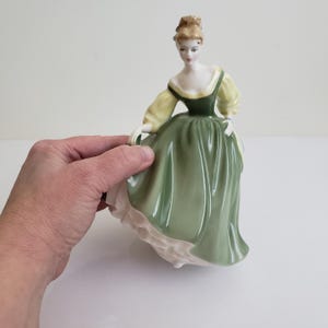May include: A porcelain figurine of a woman in a long dress. The dress is a light green colour with a white trim at the bottom. The sleeves are yellow, and the bodice is dark green. The woman has blonde hair and is holding the dress.