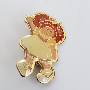 Puede incluir: Un pin de tono dorado con una imagen de dibujos animados de una niña pelirroja con un vestido amarillo y zapatos blancos. La niña sonríe y tiene el brazo levantado en el aire.