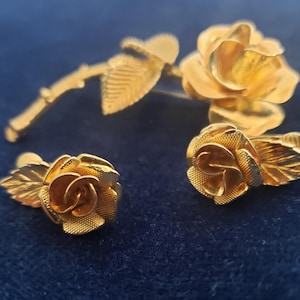 Peut inclure: Un ensemble de bijoux de couleur or avec un motif de rose. L'ensemble comprend une broche avec une tige et des feuilles, et deux boucles d'oreilles assorties. Les pétales et les feuilles de rose ont des textures détaillées. Les bijoux sont présentés sur une surface bleu foncé.