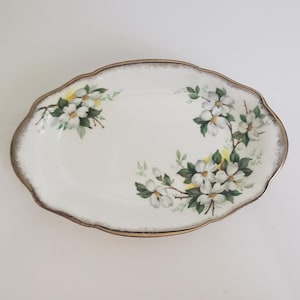 May include: Oval white serving platter with a gold rim and floral pattern. The dish has a scalloped edge and is adorned with white flowers, green leaves, and yellow highlights. A classic piece for serving starters or desserts.