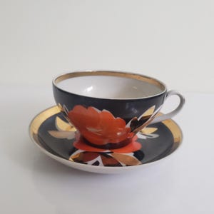 May include: A black teacup and saucer set with a floral design. The teacup and saucer have a gold rim and a red and gold floral pattern. The teacup has a white interior and a small handle. The saucer is round and has a matching design.