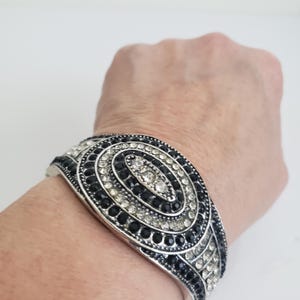 Könnte beinhalten: Ein silbernes Armreif-Armband mit schwarzen und weißen Strasssteinen, die in einem wirbelnden Muster angeordnet sind.