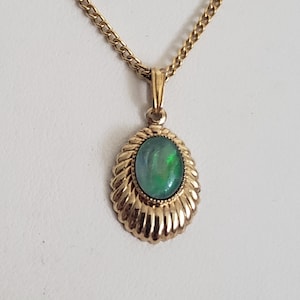 May include: A gold-toned necklace featuring an oval pendant with a shimmering green gemstone. The pendant is encased in a decorative, ridged setting. The chain is a classic curb link style, creating a timeless jewelry piece.