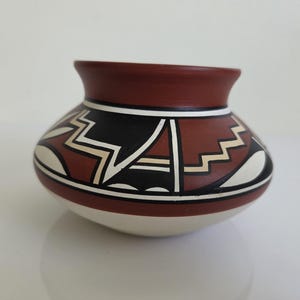 Op de afbeelding: Een kleine, ronde keramische pot met een brede, uitlopende rand en een taps toelopende voet. De buitenkant is versierd met een geometrisch patroon in zwart, wit en terracotta rood, inclusief scherpe zigzaglijnen en gebogen vormen.