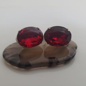 Könnte beinhalten: Ein Paar ovale Ohrringe mit tiefroten Edelsteinen und goldfarbenen Fassungen. Die Ohrringe sind auf einer braun-grau marmorierten Basis platziert. Die Edelsteine haben einen Facettenschliff, der ihr Funkeln verstärkt.