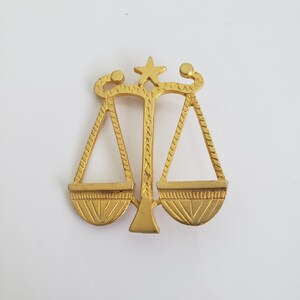May include: A gold-coloured scale of justice pendant. The pendant features a star at the top and detailed designs on the scale pans. The scales are a symbol of law and fairness.
