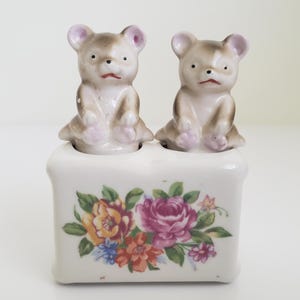 Puede incluir: Un juego de salero y pimentero de cerámica en forma de oso. Los osos son marrones y blancos con narices rosas y están sentados sobre una base blanca con un diseño floral.
