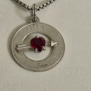 May include: A silver-toned pendant necklace featuring a circular charm with the word "JULY" and a red gemstone. An arrow pierces the center of the charm. The name "Ann" is inscribed below the arrow. The necklace chain is a silver box chain.