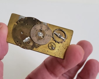 May include: A rectangular, brass-colored money clip with exposed watch gears and components. The clip features several circular gears, including one with the days of the week. The gears are various sizes and are arranged on the clip.