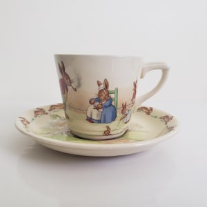以下が含まれることがあります： 牧歌的なシーンでウサギのイラストが描かれた、クリーム色のティーカップとソーサーのセット。ティーカップには取っ手があり、ソーサーは丸いです。イラストには、パイプを吸っているウサギなど、服を着たウサギが含まれています。