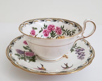 Può includere: Una tazza da tè e un piattino bianchi con un motivo floreale. La tazza e il piattino hanno un bordo dorato e un bordo nero e dorato. Il motivo floreale presenta fiori rosa, viola e gialli.