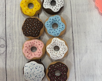 Puede incluir: Ocho donuts de fieltro coloridos con diferentes glaseados y chispas. Los donuts presentan glaseados amarillos, blancos, azules, rosas y marrones, con detalles de chispas contrastantes. Están dispuestos sobre una superficie de madera clara.