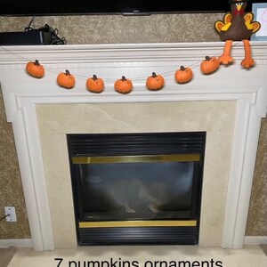 May include: A white fireplace mantel with a garland of seven orange felt pumpkins. A brown felt turkey with a black hat is perched on top of the mantel.