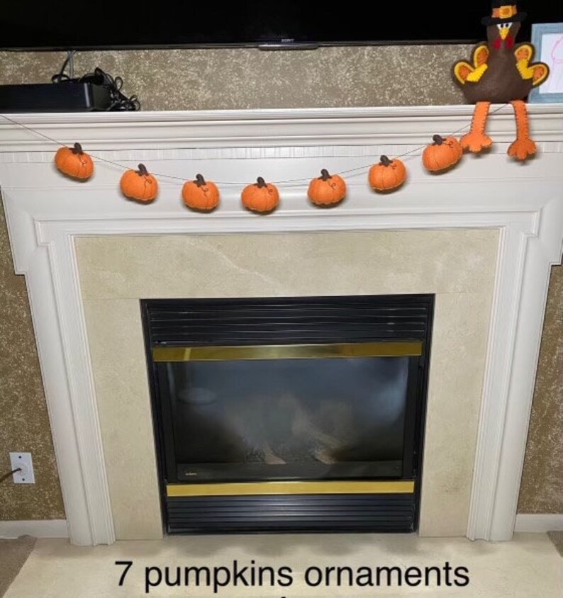 May include: A white fireplace mantel with a garland of seven orange felt pumpkins. A brown felt turkey with a black hat is perched on top of the mantel.