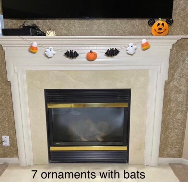 May include: A Halloween garland with seven felt ornaments, including bats, pumpkins, ghosts, and candy corn. The garland is hanging above a fireplace.