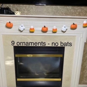 May include: A Halloween garland with nine felt ornaments, including pumpkins, candy corn, and ghosts. The garland is hanging above a fireplace with a black and gold firebox. The text "9 ornaments - no bats" is written below the garland.