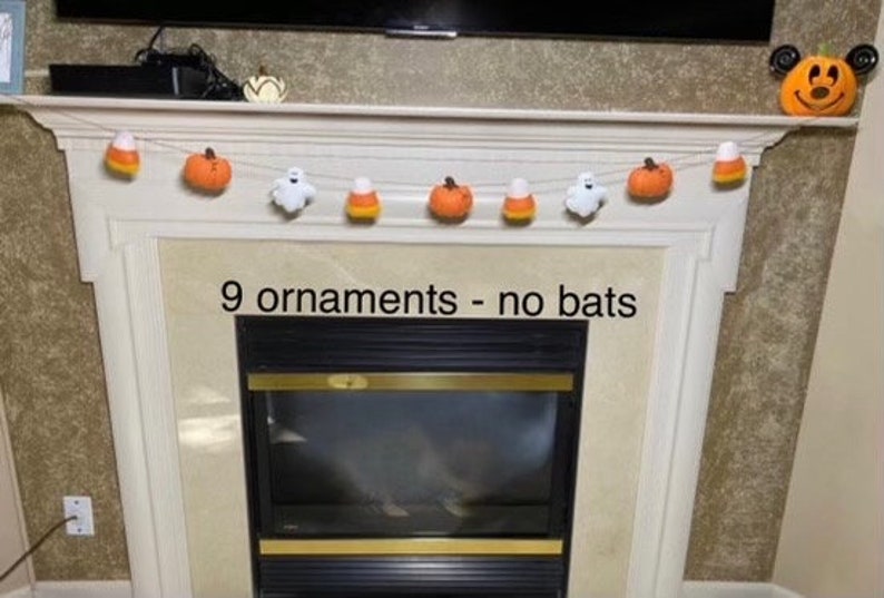 May include: A Halloween garland with nine felt ornaments, including pumpkins, candy corn, and ghosts. The garland is hanging above a fireplace with a black and gold firebox. The text "9 ornaments - no bats" is written below the garland.